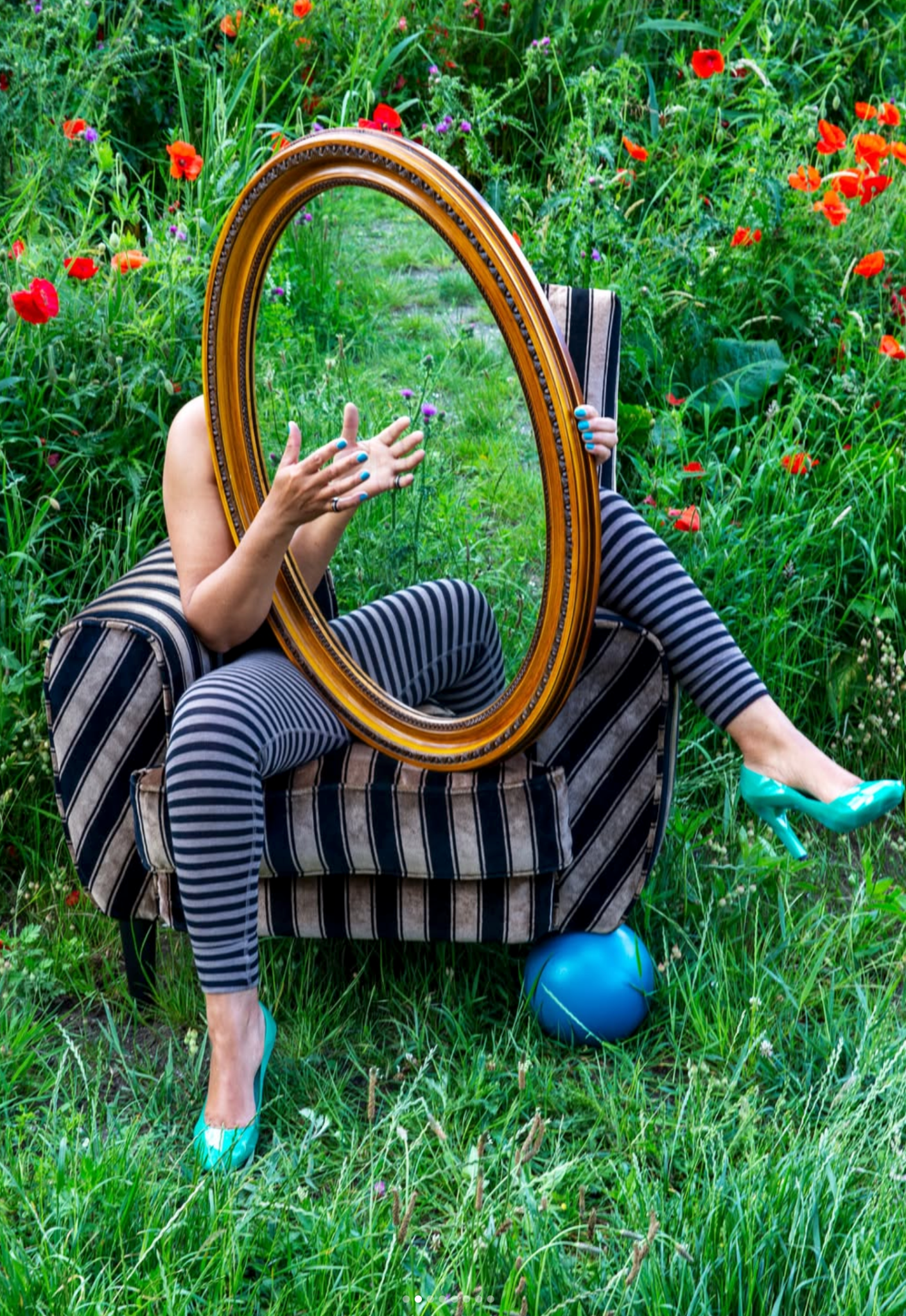 Patrícia seated in a striped chair in a wild garden, holding an oval mirror.